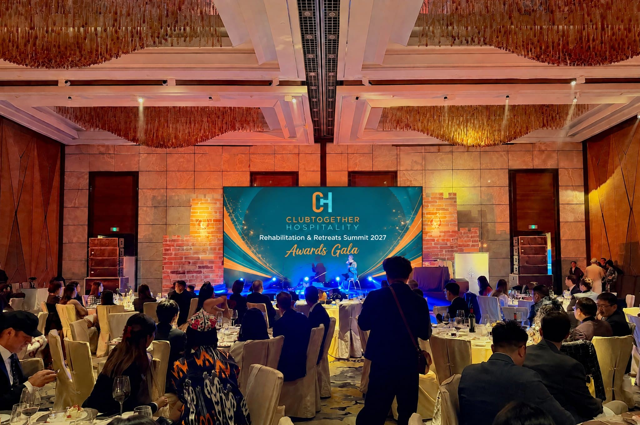 Guests at dinner tables in a ballroom, with the Awards Gala shown on the stage screen