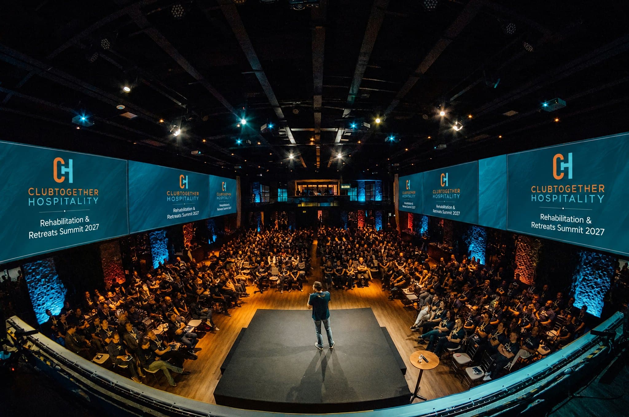 A speaker on stage in front of a full hall, with the summit shown on the big screens